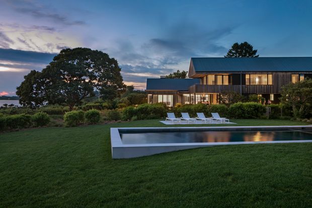 Twilight exterior view of a large contemporary two-storey home set in landscaped grounds. The building has dark vertical cladding, extensive warm-lit glazing, a pitched metal roof, and a long linear form. A minimalist rectangular reflecting or swimming pool with a pale concrete edge occupies the foreground, surrounded by manicured lawn, hedges, and established trees.