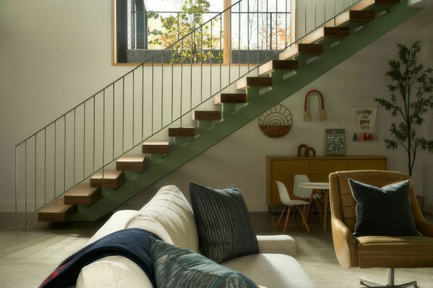 A warm, contemporary living room with a floating timber staircase and slim metal balustrade running diagonally across the space. The room includes a cream sofa, brown leather armchair, small dining or work table, built-in timber cabinetry, indoor plants, and decorative wall objects. A large window brings in natural light, while the staircase structure and green-painted stringer form a strong architectural feature.