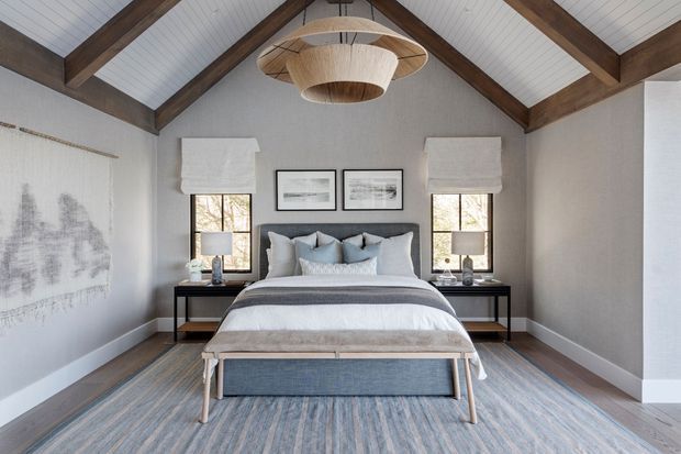 A serene, symmetrical bedroom with a centered upholstered bed, bedside tables, framed artwork, and a woven pendant light. The room has a vaulted ceiling with exposed dark timber beams, white-painted ceiling boards, pale grey walls, timber flooring, and a large muted blue-grey rug. Tall windows with light Roman blinds provide natural light, while a timber-framed bench sits at the foot of the bed.