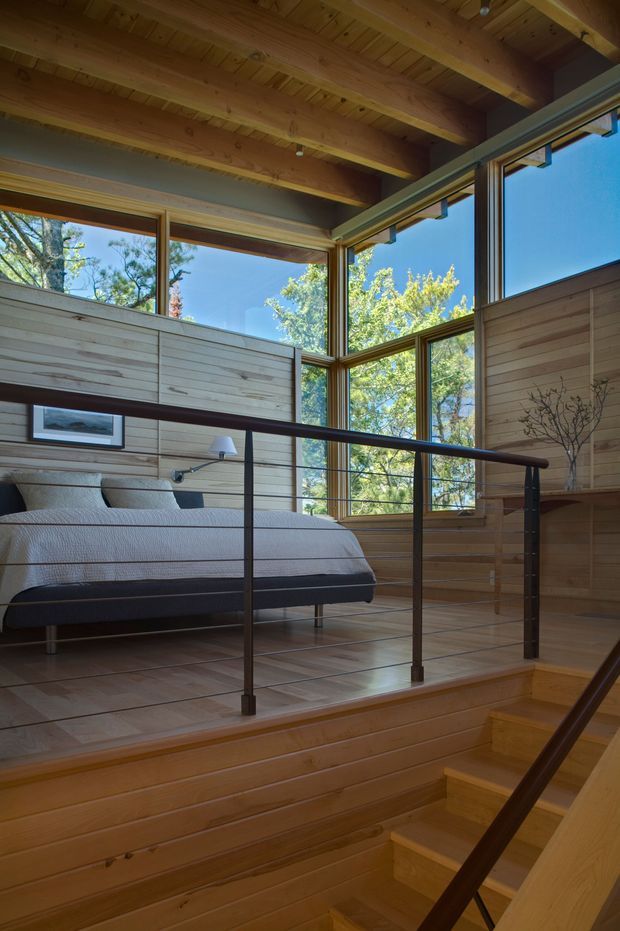 A warm, timber-lined bedroom overlooking an internal stairwell, with a low-profile bed, pale bedding, built-in wood wall surfaces, and a slender dark metal balustrade. Large corner windows bring in abundant daylight and views of leafy trees. The exposed timber ceiling beams and extensive glazing create a natural, elevated retreat-like atmosphere.