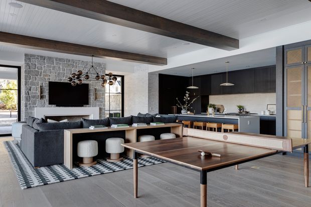 Open-plan contemporary living, dining, kitchen and recreational space with a dark timber-beamed white ceiling, exposed grey stone fireplace wall, charcoal built-in kitchen cabinetry, island seating, large sectional sofa and a table-tennis table in the foreground. Black-framed glazing and a patterned rug add contrast to the light grey floor and neutral palette.