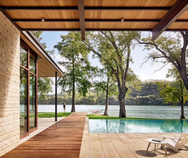 A contemporary lakeside outdoor living area viewed from beneath a timber-lined covered terrace. The scene features a pale textured masonry wall with large timber-framed glazing, a broad timber deck, a tiled pool terrace with lounge chairs, and an infinity-edge pool overlooking a lake and wooded hills. Dark steel posts and beams support the roof, while mature trees frame the water view.