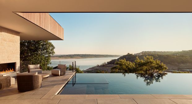 Contemporary waterfront residence viewed from an expansive outdoor terrace. A covered living area with a built-in fireplace opens onto a furnished deck and a serene infinity-edge pool, overlooking a broad coastal or lakeside inlet and wooded hills. The composition emphasizes clean horizontal lines, generous glazing/opening, warm timber soffit detailing, stone or concrete paving, and an indoor-outdoor connection.