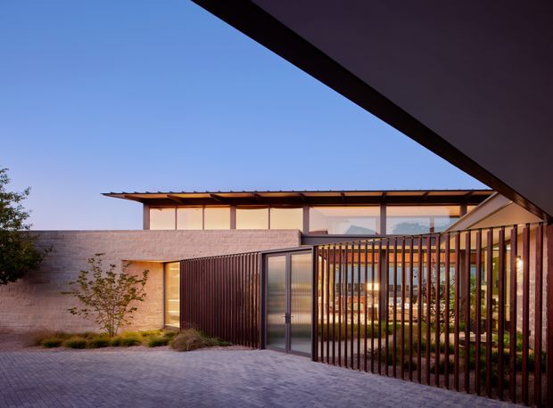 Exterior architectural photograph of a contemporary single-storey residence viewed from a paved courtyard or driveway. The composition features a low horizontal building with a shallow projecting roof, pale masonry walls, extensive glazing, dark vertical metal screening, and a gated entry. Warm interior lighting is visible through the screens and glass, while landscaped planting softens the courtyard edges.