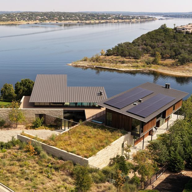 Aerial view of a contemporary multi-wing residence set on a wooded, sloping site beside a broad river or reservoir. The home has low-pitched metal roofs, extensive dark-framed glazing, timber-clad walls, masonry walls, and large rooftop solar-panel arrays. Native-looking landscaping and a planted roof terrace blend the house into the terrain, with water and a developed shoreline visible beyond.