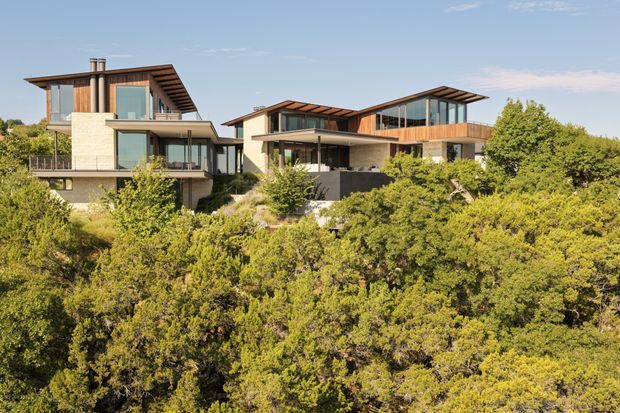 Exterior view of two contemporary multi-level residential buildings set into a densely vegetated, sloping landscape. The homes feature broad overhanging mono-pitched roof planes, extensive floor-to-ceiling glazing, warm timber cladding and soffits, pale masonry walls, dark lower-level elements, and projecting balconies or terraces. The image is photographed from across the surrounding vegetation under a clear sky.