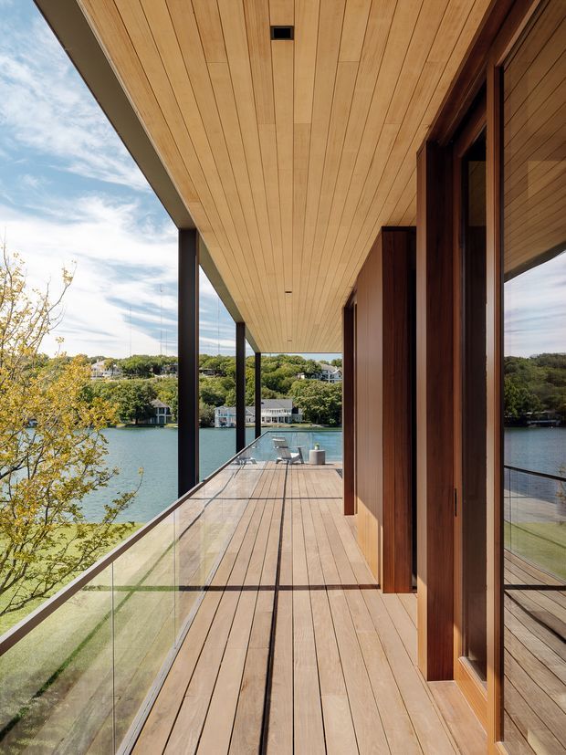 A covered waterfront balcony or terrace extending along a modern timber-clad home. The deck has warm timber boards, frameless glass balustrades, dark slender structural posts, recessed ceiling lights, and timber soffit lining. Large glazed doors and timber wall cladding run along the right side, with a swimming pool and lake visible beyond. The setting is bright, calm, and contemporary.