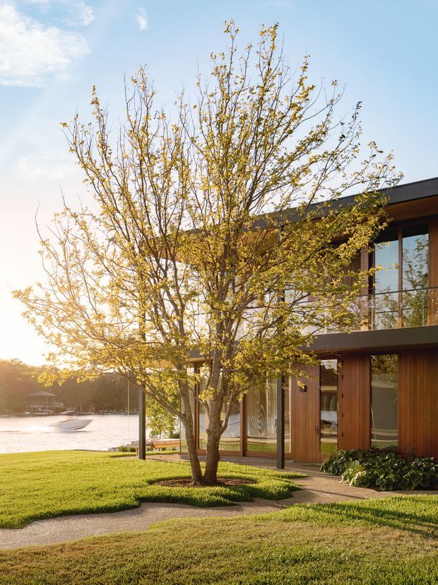 A contemporary two-storey timber-clad house beside a calm body of water, photographed in warm late-afternoon light. The home features a low-pitched dark roof, extensive floor-to-ceiling glazing, timber-framed doors and windows, a glass-balustraded upper-level balcony, and a landscaped lawn with paved paths and mature trees.