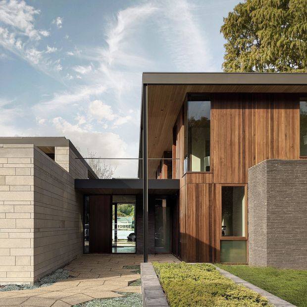 Exterior view of a refined contemporary residence with a low, flat roof and strong horizontal composition. The facade combines warm vertical timber cladding, pale horizontal masonry blockwork, dark metal framing, and expansive glazed openings. A covered entry passage connects the masonry wing to the timber-clad main volume, with geometric paving, gravel, lawn, and clipped planting in the foreground.