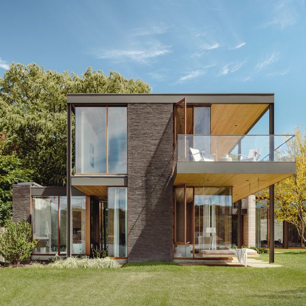 Exterior architectural photograph of a contemporary two-storey residence with a rectilinear flat-roof form, extensive floor-to-ceiling glazing, dark textured masonry cladding, warm timber soffits, and a projecting upper-level balcony with clear glass balustrade. The house is set within a landscaped suburban garden and framed by mature trees under a clear blue sky.