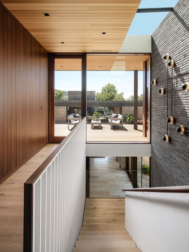 A bright, contemporary stairway and landing with a strong indoor-outdoor connection. Warm timber wall panelling, timber ceiling boards and timber flooring frame a central stair with white vertical balustrades and dark timber handrails. Full-height glazing opens toward a timber deck with outdoor seating, while a dark textured brick feature wall with sculptural wall lights runs alongside the stair. The space has abundant daylight and a double-height, open-plan feel.