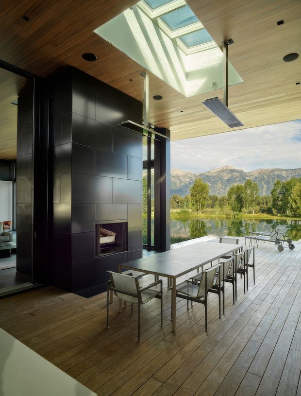 A contemporary open-plan dining and entertaining space with a long dining table beside expansive openings to a timber deck. The room features a dramatic black tiled fireplace or chimney wall, warm timber flooring and a timber-lined ceiling, with large skylights bringing in natural light. A calm lake and mountain landscape is visible beyond the deck, and slim radiant heating panels are suspended from the ceiling.
