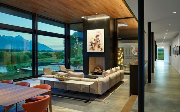 Open-plan contemporary living and dining area with expansive floor-to-ceiling glazing overlooking a dramatic mountain landscape. The room features a large sectional sofa arranged around a central fireplace and artwork, a timber-lined ceiling, polished concrete flooring, dark-framed glazing, and a timber dining table with orange upholstered chairs. A long hallway extends to the right.