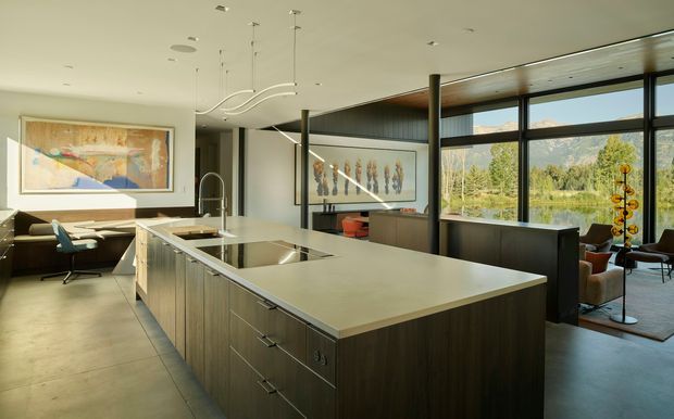 A spacious open-plan contemporary kitchen with a large central island, integrated sink and cooktop, dark timber cabinetry, and a pale stone benchtop. Floor-to-ceiling black-framed glazing opens views toward a mountainous landscape, while an adjacent dining and living area with artwork and sculptural pendant lighting is visible beyond.