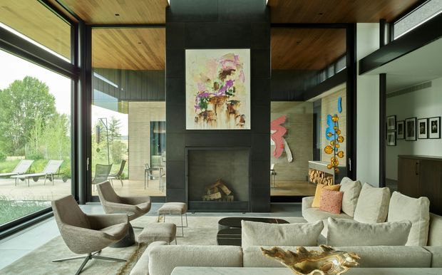 A spacious contemporary living room centred on a dark stone or concrete fireplace feature wall, with floor-to-ceiling glazing, exposed warm timber ceilings, and a large light neutral sectional sofa. Sculptural lounge chairs, ottomans, artwork, and a glimpse of an outdoor terrace create an airy, refined interior with strong indoor-outdoor connection.