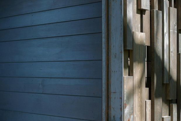 Close-up architectural detail showing dark blue-grey horizontal timber weatherboards beside a weathered vertical timber screen or cladding element with staggered projecting boards. Strong directional sunlight creates deep shadows and highlights the contrasting horizontal and vertical wood textures.