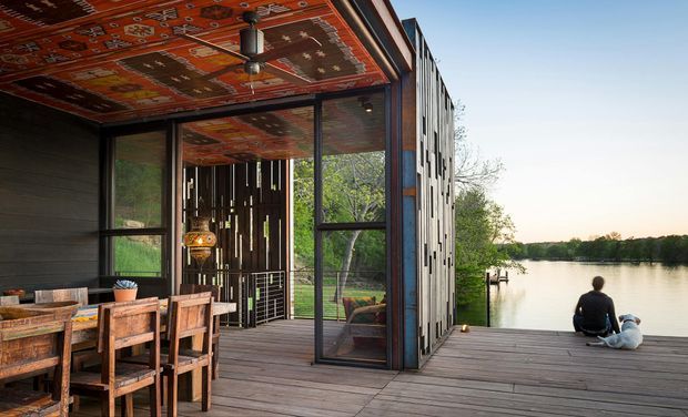 A modern waterfront pavilion or home extension opens onto a broad timber deck beside a calm lake. The covered outdoor dining area has large black-framed glazed panels, a richly patterned red and orange decorative ceiling, timber dining furniture, and a sculptural pendant light. The deck continues to the water’s edge, with a person seated overlooking the lake. The building has a narrow contemporary vertical form and dark cladding.