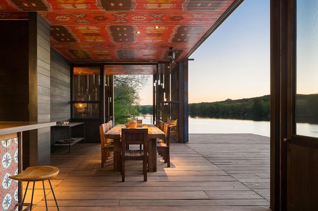 A covered timber dining terrace opens directly onto a calm waterway, with a long wooden dining table and chairs beneath a richly patterned red decorative ceiling. Dark timber-framed sliding or folding glazing, built-in cabinetry, and a patterned tiled feature panel create a warm, rustic architectural setting. The broad timber deck extends toward the water and surrounding tree-lined landscape.