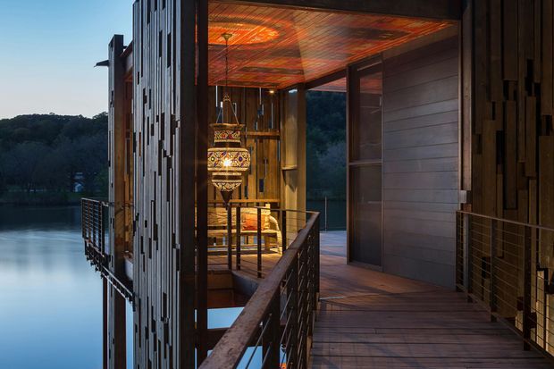 A warmly lit elevated exterior walkway or balcony beside a modern timber-clad building, overlooking a calm body of water and wooded landscape. The image features dark timber wall cladding, a richly patterned timber soffit, timber decking, metal cable balustrades, and a decorative hanging lantern visible through the open side of the structure.