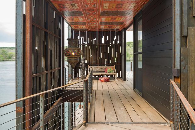 A covered elevated timber walkway or balcony running along the side of a building above a body of water. The space features weathered vertical timber screening, dark horizontal wall cladding, broad timber floorboards, cable-railing with metal posts, and a richly patterned red and orange decorative ceiling. An ornate hanging lantern and bench add visual interest, while water and landscape are visible beyond the open sides.