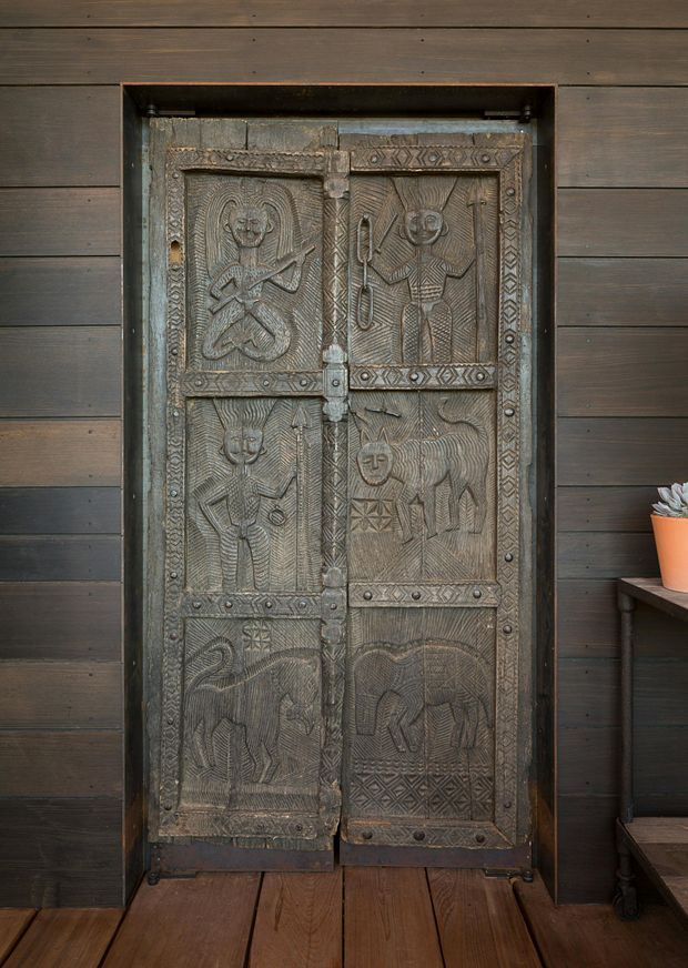 A close-up interior view of an ornate, heavily carved double wooden door set into dark horizontal timber wall panelling. The door features six decorated panels with stylised human and animal figures, geometric borders, metal hinges and fittings, and a dark aged finish. Timber flooring and a small side table with a potted plant are visible at the edges. The strong handcrafted detailing makes the door the dominant architectural feature.