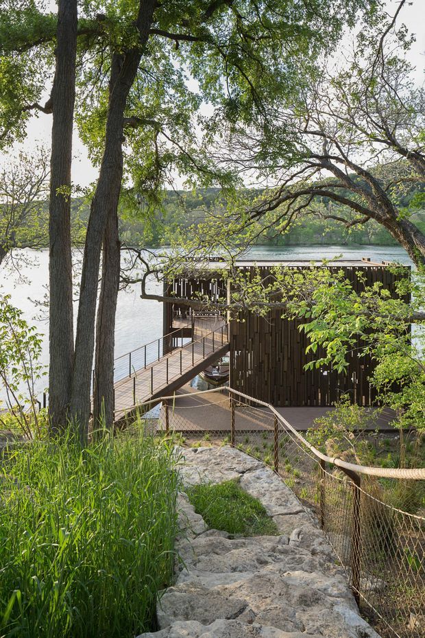 A wooded lakeside setting with a contemporary dark timber-clad structure built along the water. A rocky pedestrian path and rope-and-mesh railing descend through tall grasses and mature trees to an elevated timber boardwalk and deck beside the building. The lake and wooded opposite shore are visible beyond.