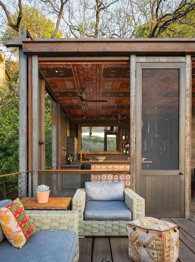 A compact covered outdoor entertaining space with a rustic industrial enclosure, weathered steel framing, timber decking, woven lounge furniture, and a richly patterned decorative ceiling. The space includes a bar area with tiled cabinetry, mirror, shelving, and pendant lighting, accessed through a glazed timber-framed door. Colourful patterned cushions and an upholstered ottoman add eclectic character, while surrounding trees create a garden setting.