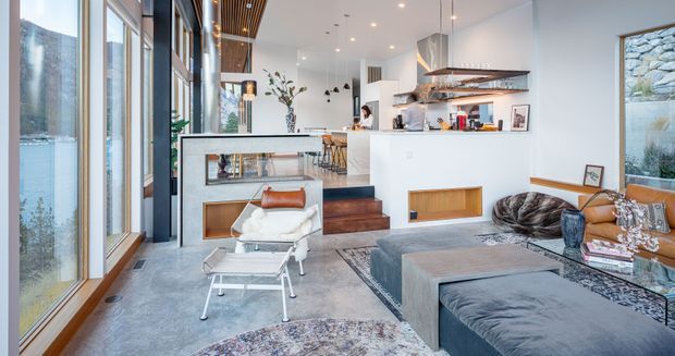Open-plan contemporary living and kitchen space with full-height glazing, a white kitchen with island seating, integrated timber cabinetry, a sunken living area, and a large grey sectional sofa. Warm timber ceiling detailing, pendant lighting, neutral finishes, and a visible stone-clad exterior wall create a refined, light-filled interior.