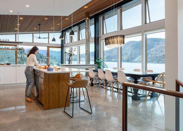 A spacious, open-plan contemporary kitchen and dining area with expansive floor-to-ceiling glazing overlooking a forested mountain and lakeside landscape. The kitchen features a wood-clad island with bar seating, white cabinetry, pendant lights, polished concrete flooring, and a slatted timber ceiling. A long dining table sits beside the glazing, with a staircase and glass balustrade in the foreground.