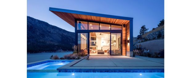 A contemporary single-storey pavilion-style house beside a swimming pool and mountain lake landscape at dusk. The building has extensive floor-to-ceiling glazing, a strongly projecting flat roof, dark blue exterior framing, and warm timber soffit and wall cladding. The illuminated interior appears to contain an open-plan living area, while the foreground is dominated by the pool terrace.