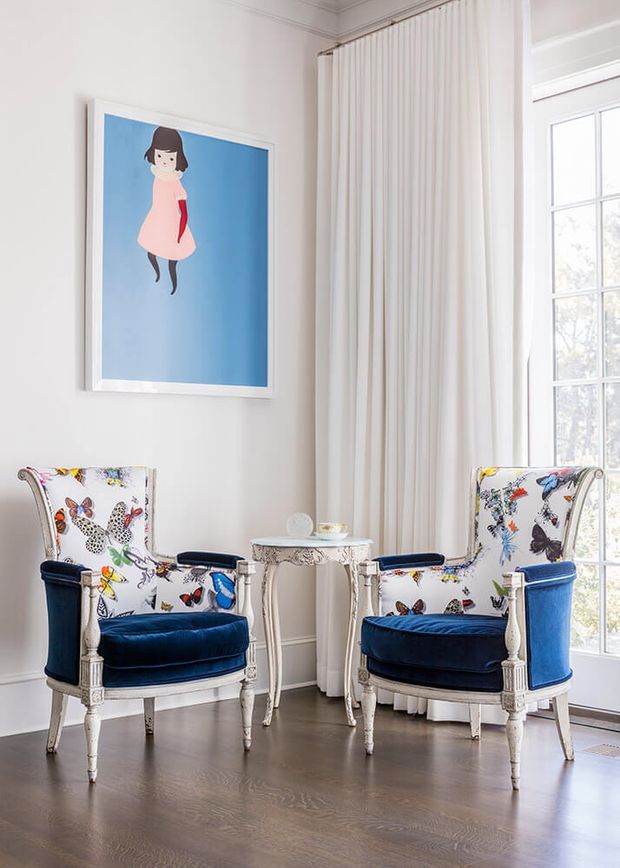 A bright, elegant sitting area with two ornate upholstered armchairs facing a small decorative round table. The chairs feature white fabric with colourful butterfly and floral motifs, deep navy velvet seats and arm panels, and carved white-painted timber frames. A large blue artwork hangs on the white wall, while full-height white curtains and a tall multi-pane window bring in abundant natural light. The room has dark timber flooring and refined traditional detailing.