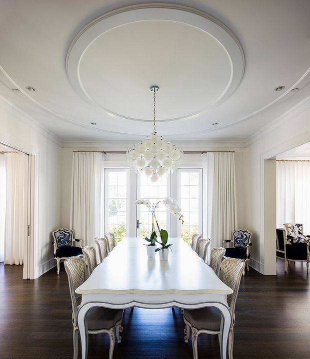 Elegant formal dining room with a long sculpted white dining table surrounded by ornate upholstered and woven-back chairs. The room features dark timber flooring, pale walls with elaborate layered ceiling mouldings and a large circular ceiling detail, tall multi-pane French doors with full-height cream curtains, and a dramatic clustered pendant light. Openings on either side reveal adjoining light-filled rooms.