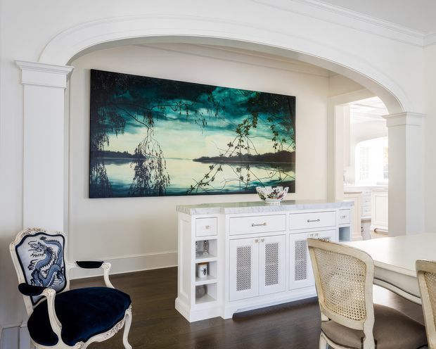 Elegant traditional dining area with a large white arched opening and decorative trim, dark timber flooring, a white built-in sideboard with patterned grille doors, and a white dining table with woven-back chairs. A large blue-green landscape artwork dominates the wall, while an ornate blue upholstered armchair adds contrast. A bright adjoining room is visible through the archway.