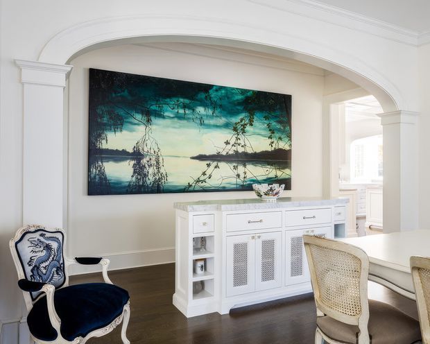 Elegant traditional dining area viewed through a wide arched opening, featuring white architectural trim and columns, a dark timber floor, a large landscape artwork, and a white built-in sideboard with decorative mesh-front doors and open shelving. Blue upholstered ornate chairs and woven dining chairs add contrast. A bright adjoining kitchen or utility space is visible through the opening on the right.
