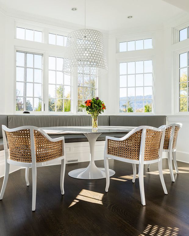 Bright dining nook with a built-in curved banquette beneath large multi-pane windows. A round white pedestal dining table is surrounded by white-framed woven cane-back chairs, with a large white lattice pendant light overhead. Dark timber flooring contrasts with the predominantly white walls, trim, ceiling, and window frames. The setting appears airy and refined, with a distant landscape visible through the glazing.