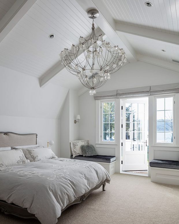Bright, elegant bedroom with a large upholstered bed, pale patterned bedding, built-in window seats, and French-style glazed double doors. The room features white beadboard walls and vaulted ceiling panels with exposed white beams, a large ornate crystal chandelier, soft grey upholstery, and light carpet. The overall design is refined, airy, and traditionally styled.