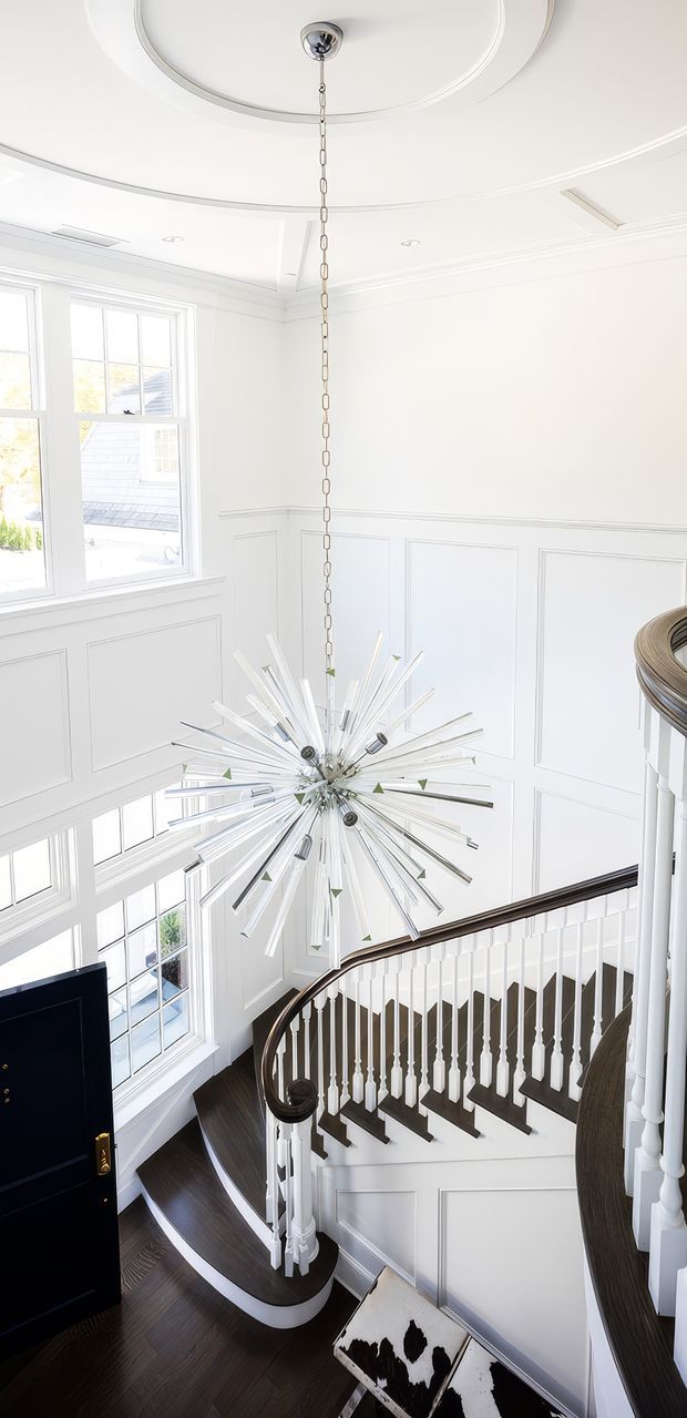 A bright, grand residential stairway and landing viewed from above, featuring a sweeping curved staircase with dark timber treads, white turned balusters, a dark polished handrail, and white paneled walls. A large sculptural starburst pendant hangs through the stair void from an ornate circular ceiling medallion. Tall multi-pane windows flood the space with natural light, while dark flooring and a black entry door provide contrast.