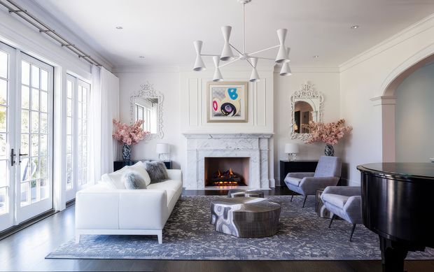 Bright, elegant living room with white walls and trim, tall French doors, a marble fireplace with a lit fire, sculptural white pendant lighting, a white sofa, upholstered armchairs, decorative mirrors, and a glossy black grand piano. The room combines classic architectural detailing with contemporary furnishings.