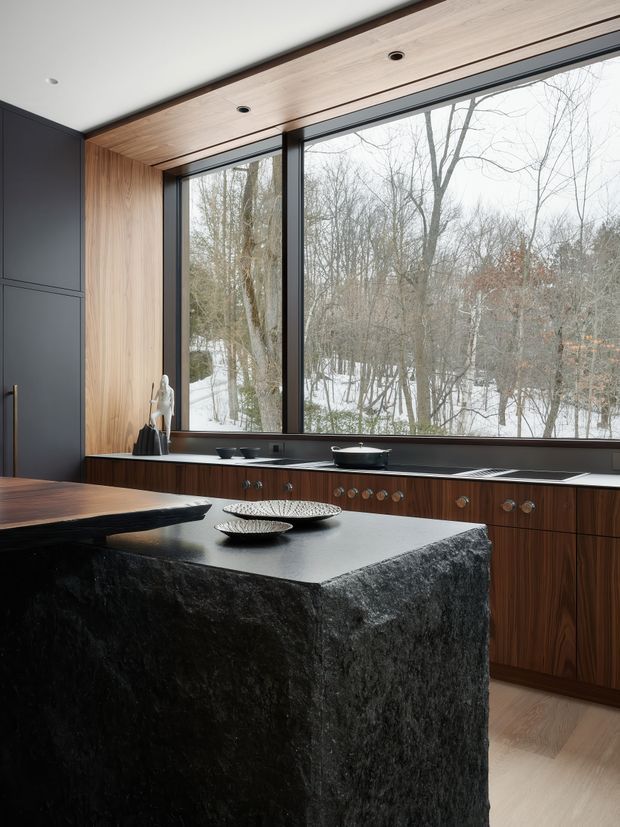 A sophisticated modern kitchen with a large dark stone island and integrated cooktop, warm timber cabinetry and ceiling lining, matte dark wall panels, and extensive black-framed windows overlooking a snowy wooded landscape. The space features understated decorative objects, recessed lighting, and a refined natural-material palette.
