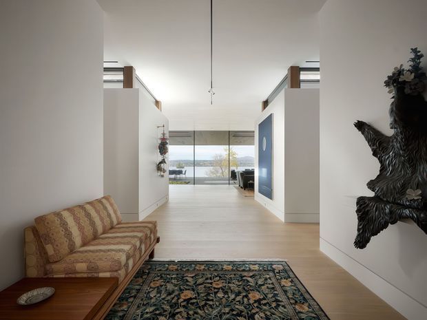 A bright, refined residential hallway or entry space opening toward full-height glazing and a waterside view. The minimalist white interior features pale timber flooring, built-in white joinery, a blue accent panel, a patterned vintage sofa, a dark wooden coffee table, an ornate area rug, and sculptural wall-mounted decor. A high, open ceiling and long sightline create an airy sense of space.