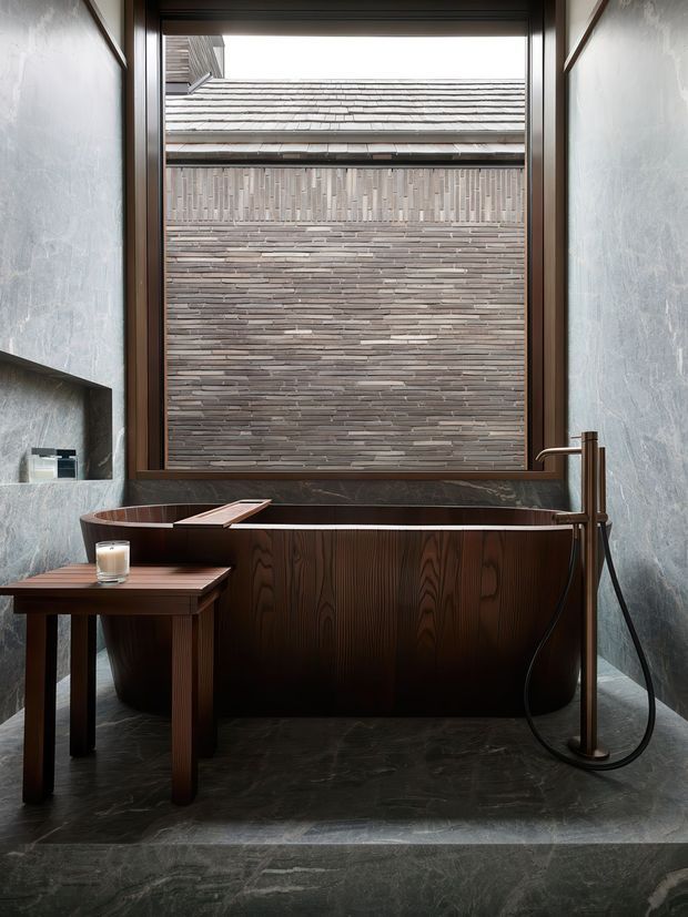 A dramatic, compact bathroom featuring a freestanding dark timber bathtub with integrated angular detailing, a floor-mounted brushed metal tap and handheld shower, and a matching timber side table. Large grey stone-textured wall panels and dark stone flooring frame a full-height window overlooking a layered brick wall and tiled roof. The scheme is moody, refined, and strongly material-focused.