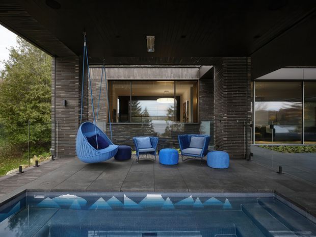Covered outdoor terrace beside a swimming pool, with dark brick walls, extensive glazing, built-in architectural lighting, and blue outdoor lounge furniture. The space appears connected to the main residence through large openings and reflective windows, with landscaping visible beyond.