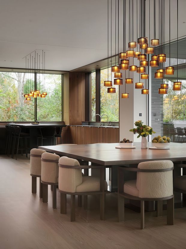 A refined open-plan dining and kitchen interior with a large rectangular dining table surrounded by upholstered dining chairs. A dramatic cluster of suspended amber-glass pendant lights hangs above the table, while a second smaller cluster illuminates the dark kitchen island in the background. The room features floor-to-ceiling glazing overlooking trees, vertical timber wall panelling and cabinetry, a pale timber floor, dark stone or composite benchtops, and a glimpse of a grey brick feature wall through the glazed opening. The overall design is warm, contemporary, and sophisticated.