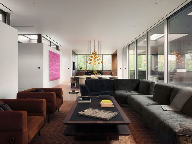 A spacious contemporary living room with a large dark sectional sofa and matching low coffee table in the foreground. Brown lounge chairs sit to the left, while a dining area and sculptural pendant light are visible beyond. Full-height glazed walls on the right provide views into an adjacent space and greenery outside. White walls, polished timber flooring, a vibrant pink artwork, and a warm patterned rug create a refined, modern interior.
