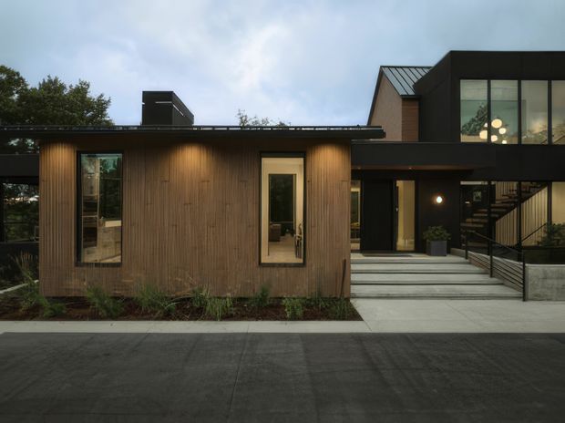 Exterior view of a contemporary multi-level residence at dusk, featuring a dark metal-clad upper volume, a projecting low flat-roof section, and a warm vertically timber-clad facade. Large glazed openings, recessed entry lighting, concrete steps, and black metal railings define the frontage.