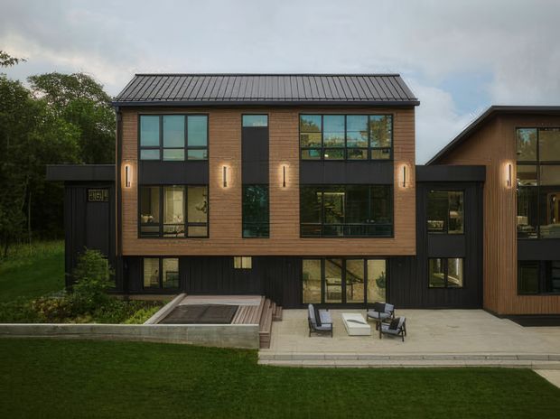 Exterior view of a contemporary multi-level residence with a strong rectangular composition, dark standing-seam metal gable roof, warm timber-toned cladding, extensive black-framed glazing, and dark vertical cladding at the lower level. A paved terrace with outdoor seating occupies the foreground, with a small sunken water or garden feature to the left. The home is set within a landscaped suburban or wooded site.