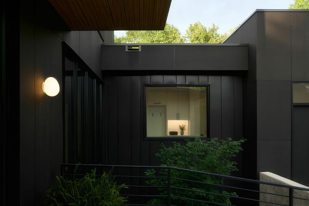 A dark, contemporary flat-roofed building facade viewed from an elevated outdoor walkway or courtyard. Black or charcoal cladding, large glazed openings, a projecting timber-lined soffit, minimalist wall lighting, and black metal railings frame a landscaped planted area. A softly lit interior room is visible through the central window.