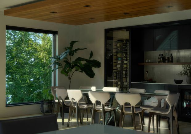 A contemporary dining and entertaining area with a long dining table surrounded by light-coloured modern chairs. The room features a large picture window overlooking leafy greenery, a warm timber-lined ceiling with recessed downlights, pale walls, and a dark built-in bar or display joinery wall with open shelving and cabinetry. Indoor plants add a natural element, while sunlight creates a calm, refined atmosphere.