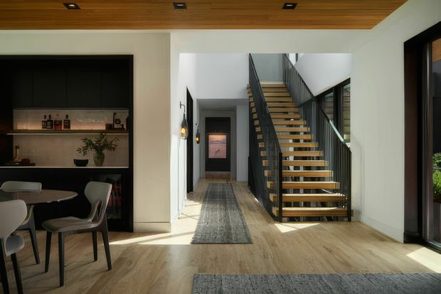 A bright contemporary open-plan interior focused on a staircase and hallway. An open-riser timber staircase with dark metal stringers and balustrades rises beside a light-filled corridor, while a dining area and dark built-in home bar or display joinery occupy the left side. The space has pale timber flooring, white walls, a timber-lined ceiling with recessed lighting, large dark-framed glazing, and neutral grey rugs.