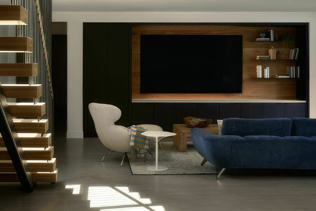 A sophisticated living room with a built-in dark timber and black media wall, integrated shelving, large television, and low cabinetry. The room features a deep blue sofa, sculptural cream lounge chair, small round side table, patterned throw, area rug, and polished grey floor. A dramatic open-riser timber staircase with dark supports is visible at the left, and sunlight creates strong geometric shadows across the floor.